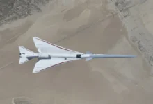 NASA’s white, blue, and red X-59 research aircraft flies over the Mojave Desert near NASA’s Armstrong Flight Research Center in Edwards, California, during its third flight. The aircraft is centered in the frame, viewed from above with the canopy, both wings, NASA and X-59 logos visible, and the landing gear down with one wheel visible. Below, rugged terrain transitions into the smooth texture of a dry lakebed as the aircraft, nearly 100 feet long, flies toward the right side of the frame.