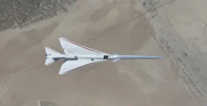 NASA’s white, blue, and red X-59 research aircraft flies over the Mojave Desert near NASA’s Armstrong Flight Research Center in Edwards, California, during its third flight. The aircraft is centered in the frame, viewed from above with the canopy, both wings, NASA and X-59 logos visible, and the landing gear down with one wheel visible. Below, rugged terrain transitions into the smooth texture of a dry lakebed as the aircraft, nearly 100 feet long, flies toward the right side of the frame.