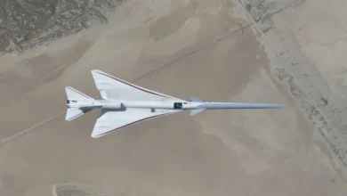 NASA’s white, blue, and red X-59 research aircraft flies over the Mojave Desert near NASA’s Armstrong Flight Research Center in Edwards, California, during its third flight. The aircraft is centered in the frame, viewed from above with the canopy, both wings, NASA and X-59 logos visible, and the landing gear down with one wheel visible. Below, rugged terrain transitions into the smooth texture of a dry lakebed as the aircraft, nearly 100 feet long, flies toward the right side of the frame.