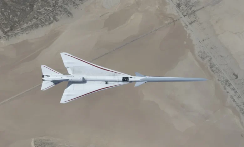 NASA’s white, blue, and red X-59 research aircraft flies over the Mojave Desert near NASA’s Armstrong Flight Research Center in Edwards, California, during its third flight. The aircraft is centered in the frame, viewed from above with the canopy, both wings, NASA and X-59 logos visible, and the landing gear down with one wheel visible. Below, rugged terrain transitions into the smooth texture of a dry lakebed as the aircraft, nearly 100 feet long, flies toward the right side of the frame.